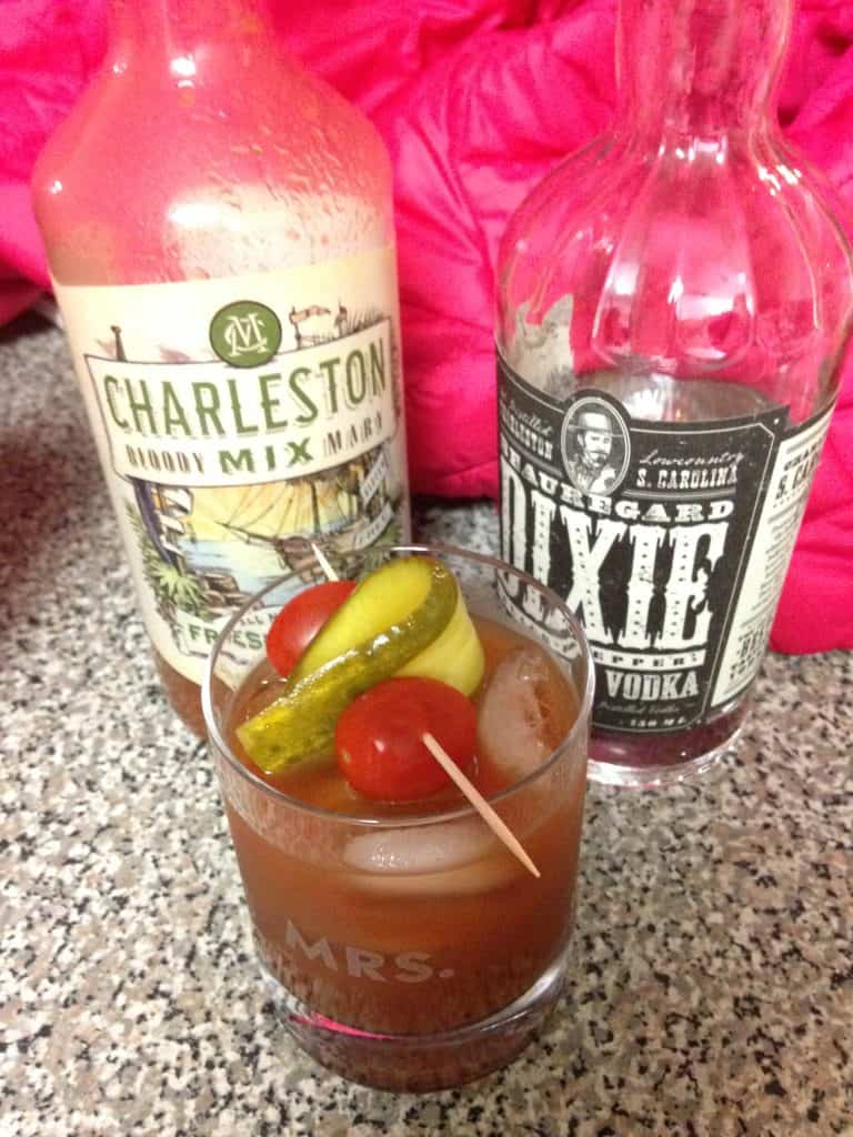 A bottle of Charleston bloody mary mix next to a bottle of Dixie vodka and a short glass with a red-colored åcocktail topped with pickles, tomatoes, and olives in this color, close-up photo for 78 Southern Phrases to Blow Your Hair Back, Cocameca.com photo © Canva
