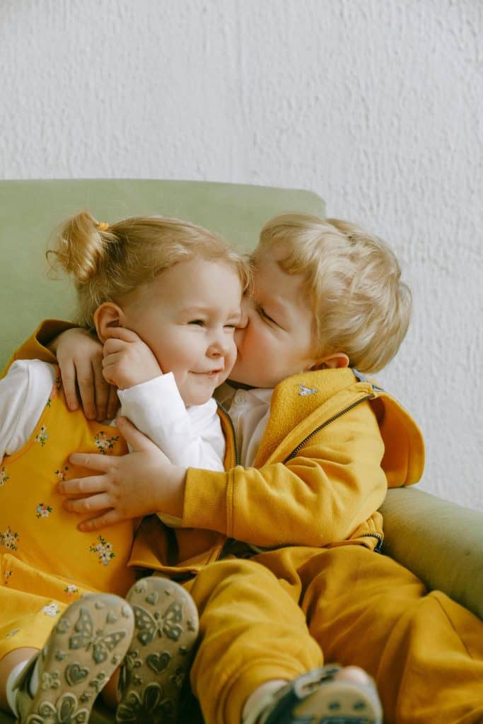 "Give me some sugar": two blonde children in ocre-colored soft clothing kiss gently on a sofa in this color, close-up photo for 78 Southern Phrases to Blow Your Hair Back, Cocameca.com photo ©Shevtsa for Pexels