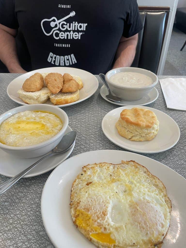 A person wearing a black t-shirt with "Guitar Center Georgia" in white screenprint faces a linoleum-topped table with plates of food - fried eggs, grits, biscuits and fried chicken at The Majestic in Atlanta, GA, photo ©Kate Dana