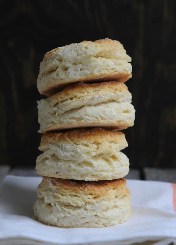 A stack of four, fluffy, buttery, layered buttermilk biscuits, golden brown with flaky insides, rest on a table in this photo ©Jodie Morgan for Unsplash