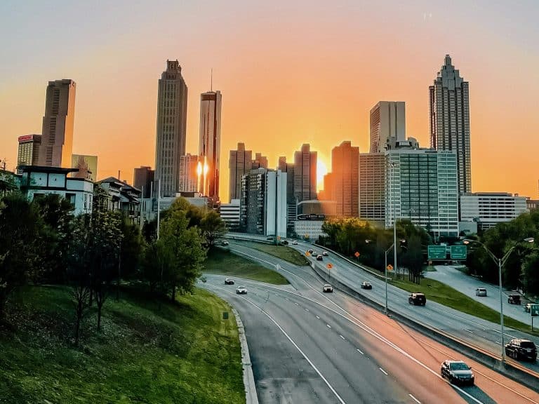 A colorful view of the skyline at daybreak shows tall buildings against a sunrise, with few cars on the expressway and green hills in the foreground, a typical scene when you explore Atlanta like a local, photo ©Marianna Smiley for Unsplash