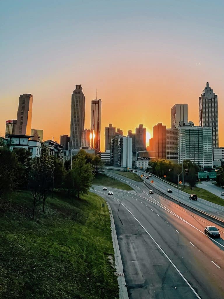 A gorgeous view of the sun rising over distant Atlanta, GA with a near-empty expressway in the foreground next to a grassy knoll in this photo ©Marianna Smiley for Unsplash