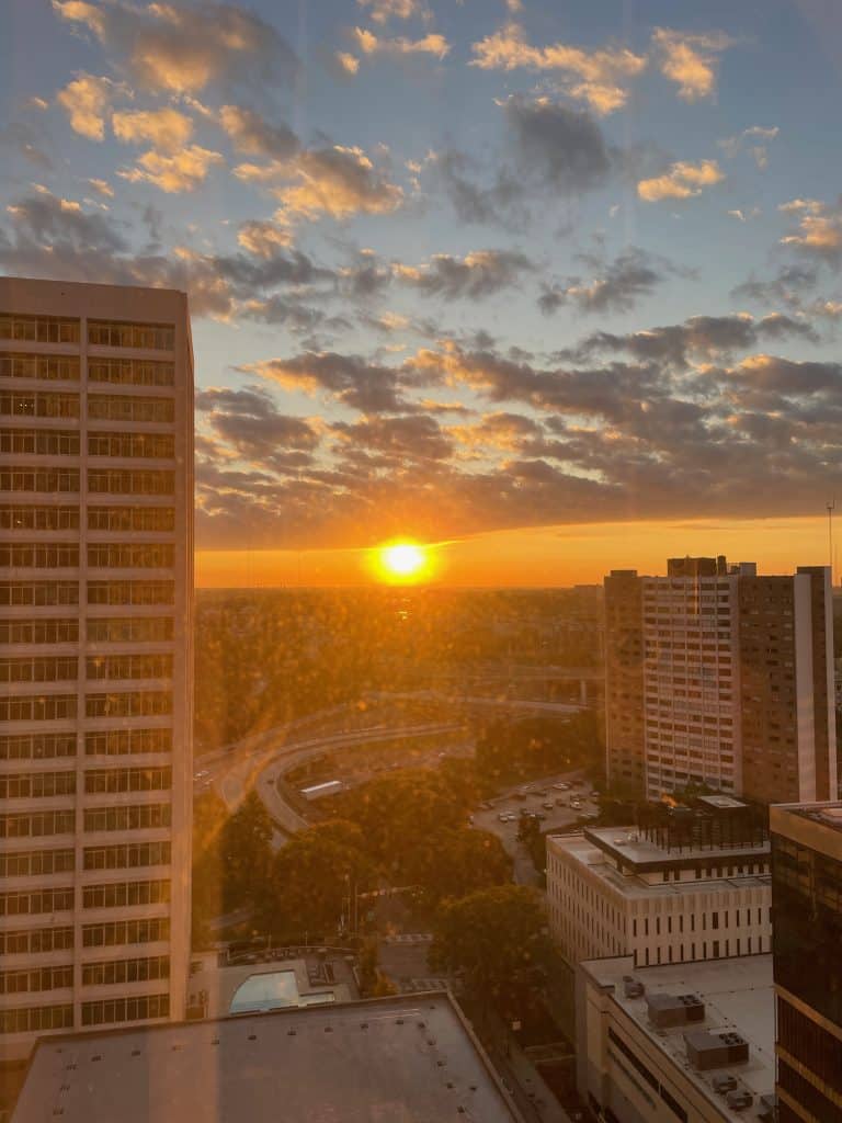 The sun rises over the city and buildings with a cloudy sky, illuminating this image taken from a room high above inside the Hilton Atlanta in Atlanta, GA, photo ©Kate Dana