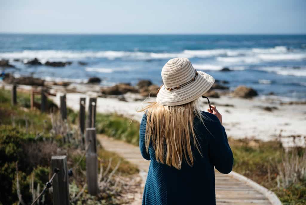 A person in a blue sweater with a white beach hat faces the ocean walking on a boardwalk in this photo by ©Vital Sinevich for Unsplash