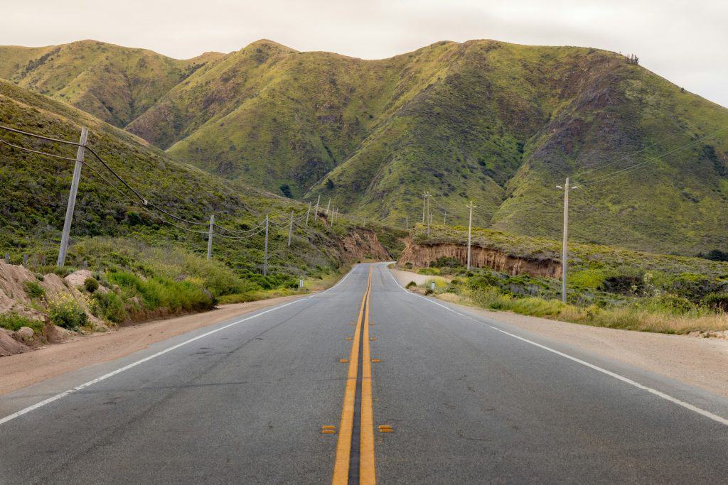 A two-laned asphalt road with a double yellow line down the middle heads into the distant, green mountains along the Monterey Peninsula near Carmel, California, photo ©Martin Adams for Unsplash