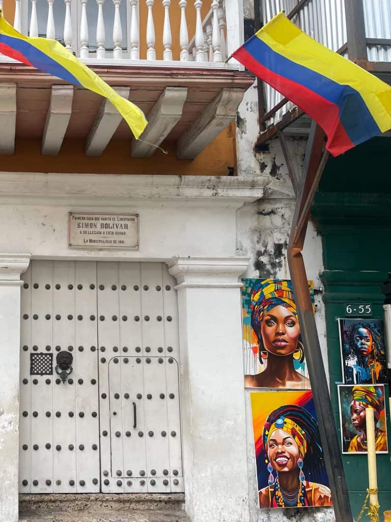 National flags wave from a second-story balcony above paintings of lovely Afro Colombian women for sale on the street below in Cartagena, Colombia, a city similar to Charleston, S.C., photo ©Kate Dana