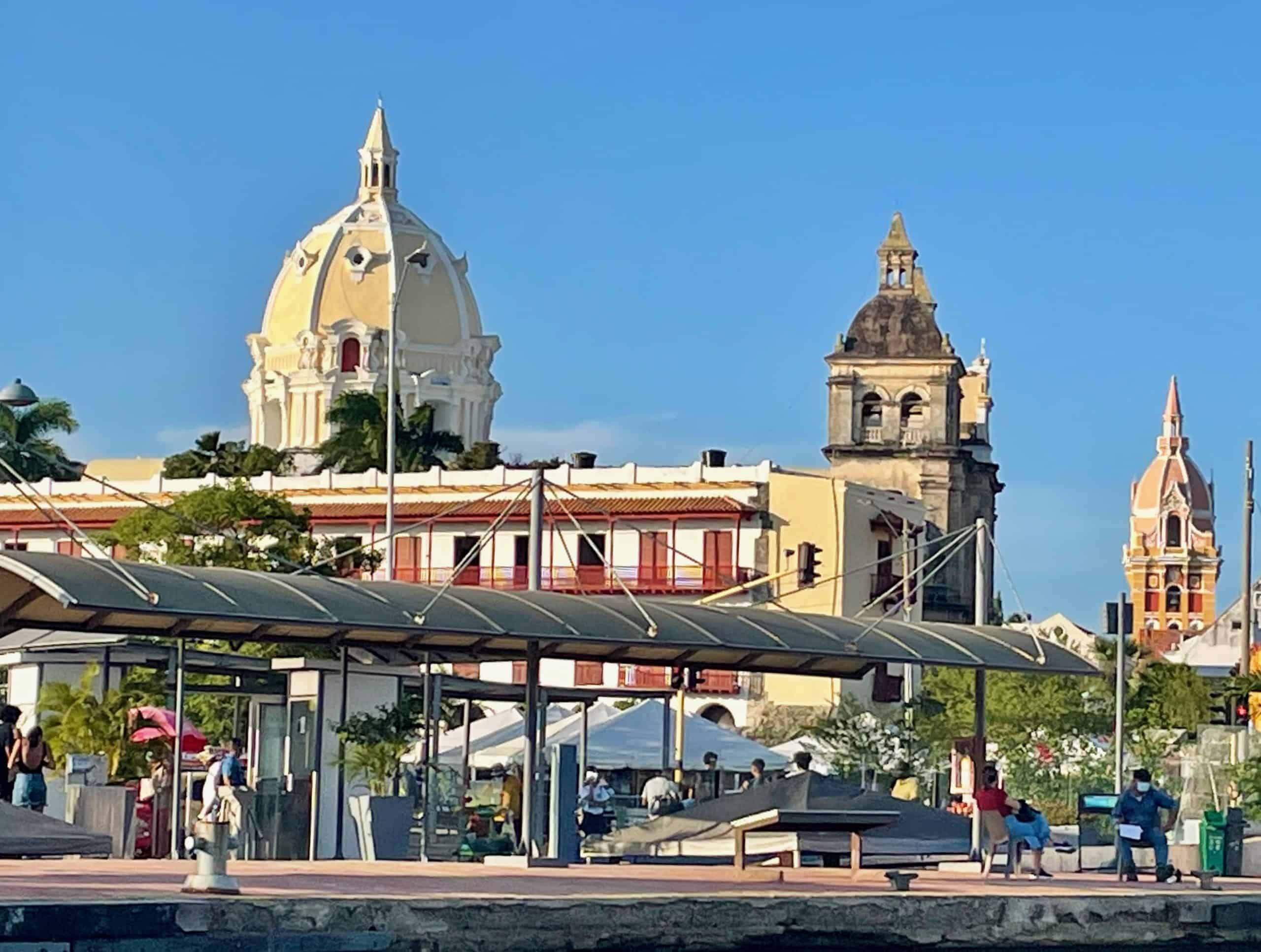 The pink and gold Spanish renaissance dome of Iglesia Santa Catalina de Alejandría and the strong towers of Catedrál San Pedro Claver rises in the background over a city street and the harbor in the foreground, in this photo taken from a boat near the Centro in Cartagena, Colombia, a city similar to Charleston, S.C., photo ©Kate Dana