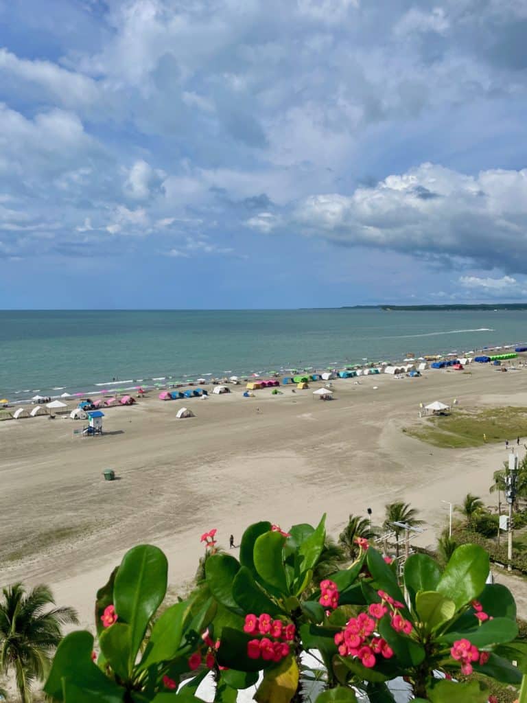 Brilliant fuschia flowers spill over the edge of a planter next to a pool overlooking the distant beach in La Boquilla with chairs and tents set up beside the water near Cartagena, Colombia, a city similar to Charleston, S.C., photo ©Kate Dana
