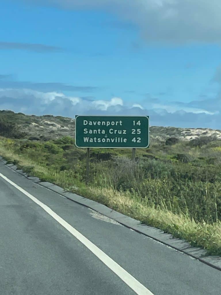 A green and white highway sign announces the driving distance to Davenport, Santa Cruz and Watsonville along a roadside with grassy fields, photo © Kate Dana