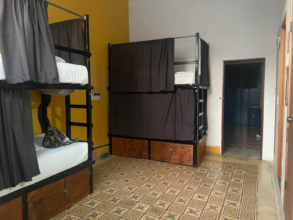 Tall dark bunk beds against a golden colored wall and white wall, with ornate clay tiles on the floor of a dormitory at Che Nomadas hostel in Merida, Mexico, photo ©Kate Dana