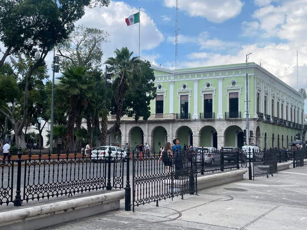A pale green historic building with arches on the balcony and the Mexican flag flying overhead against a blue sky in Merida, Mexico, photo ©Kate Dana
