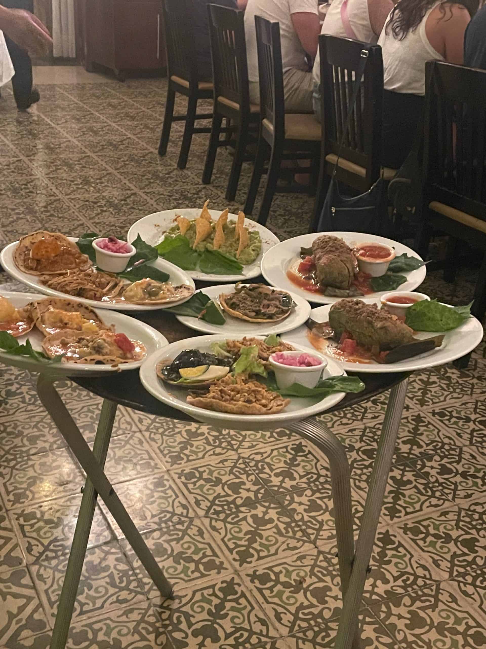 7 large dinner plates filled with Yucatán inspired traditional dishes sit on a tray stand at La Chaya Maya restaurant in Merida, Mexico, photo ©Kate Dana