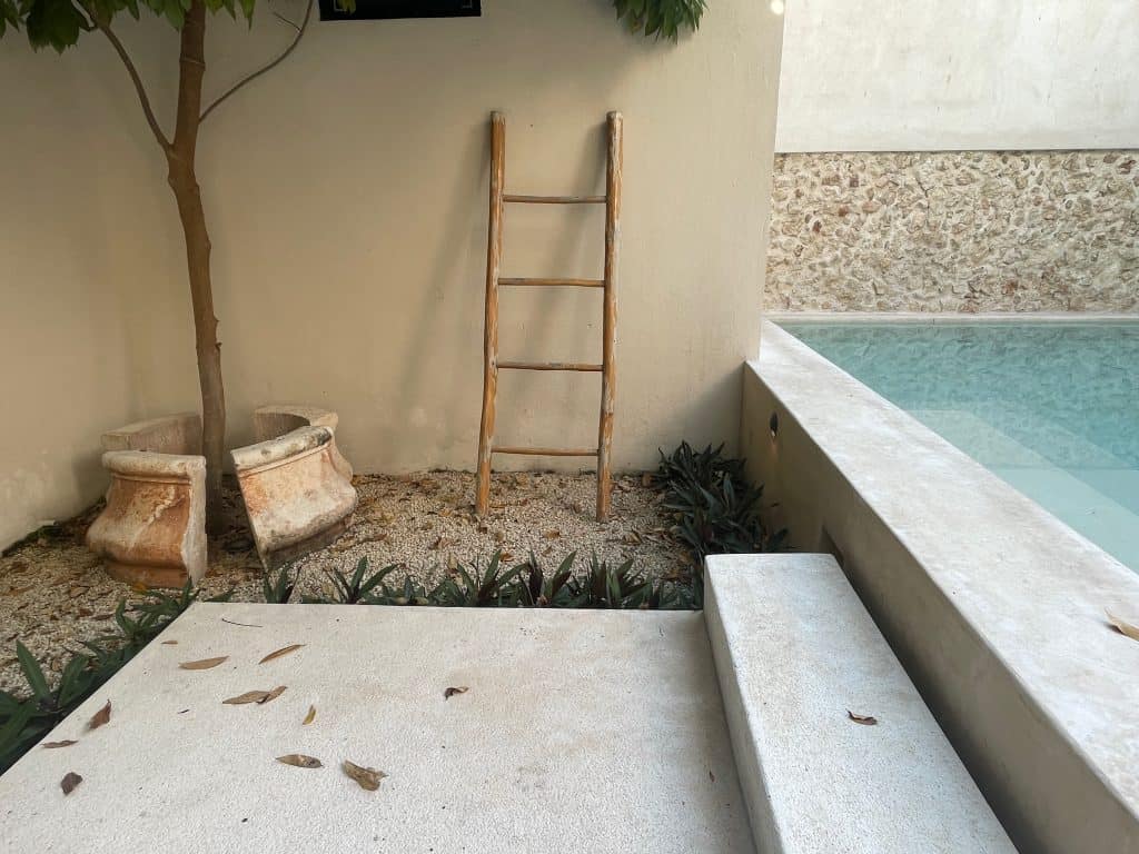 A rustic handmade ladder leans against a stucco wall next to a shimmering aqua stone pool, photo ©Kate Dana
