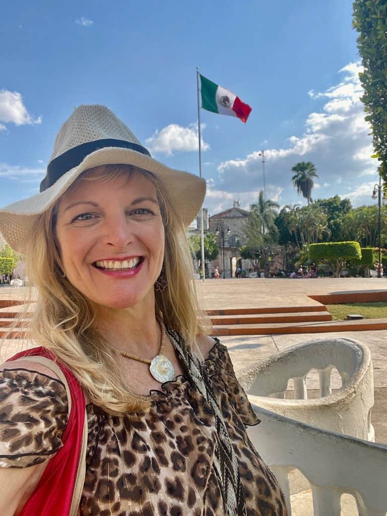 Kate Dana, founder of Cocmeca, smiles in the main plaza of Merida, Mexico with a large Mexican flag flying in the background before a sunny, blue sky, photo ©Kate Dana