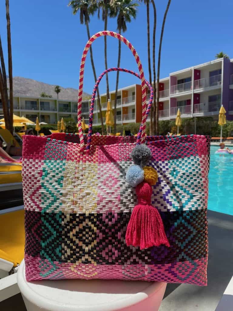 A beautiful, colorful, handmade tote bag with black, pink, blue, and yellow diamond pattern with a handmade pink, blue, yellow, and black pompom sits on a table near a pool on a sunny day in this photo adding to the article Mexican Slang for Beginners: Phrases to Know photo ©Kate Dana