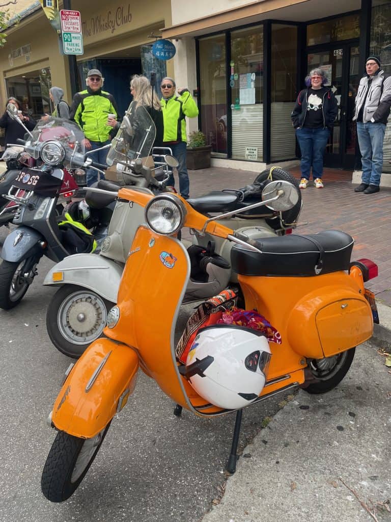 An orange, 1973 Vespa Primavera small frame scooter sits parked next to a vintage, silver Vespa Sprint 150 before some retail shops and eateries in Pacific Grove, California, photo ©Kate Dana
