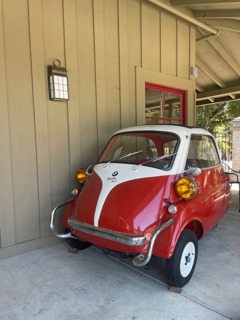 A tiny, red and white BMW Iseta car sits parked on the sidewalk outside the showroom of the Moto Talbott museum in Carmel Valley, California, photo ©Kate Dana