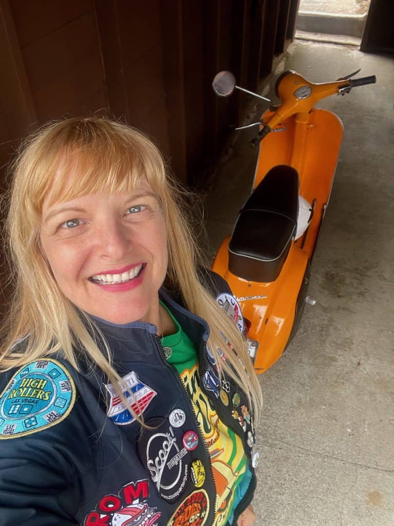 Cocameca founder Kate Dana smiles at the camera next to an orange, 1973 Vespa Primavera small frame scooter parked in the rustic carport of the Director's Cottage at Asilomar Hotel & Conference Grounds in Pacific Grove, CA, photo ©Kate Dana