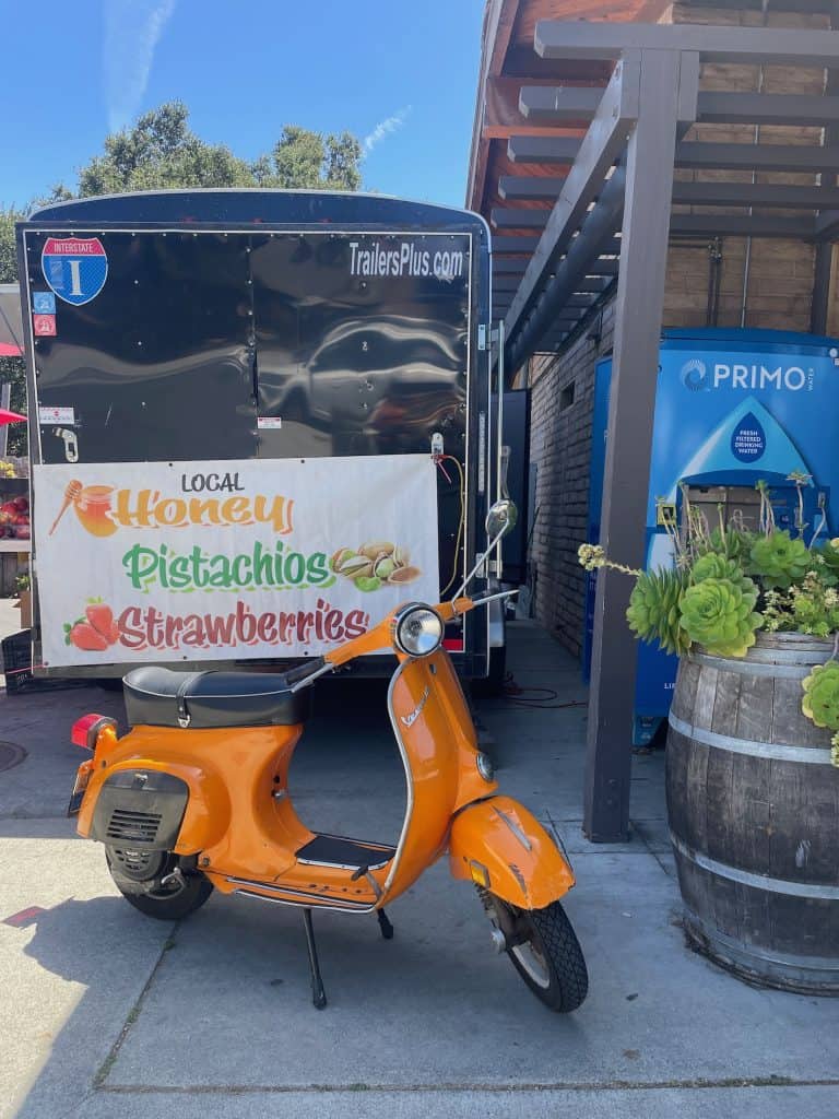 An orange, 1973 Vespa Primavera small frame scooter sits parked in front of a colorful sign offering Honey, Pistachios, and Strawberries on a black trailer in Carmel Valley, California, photo ©Kate Dana