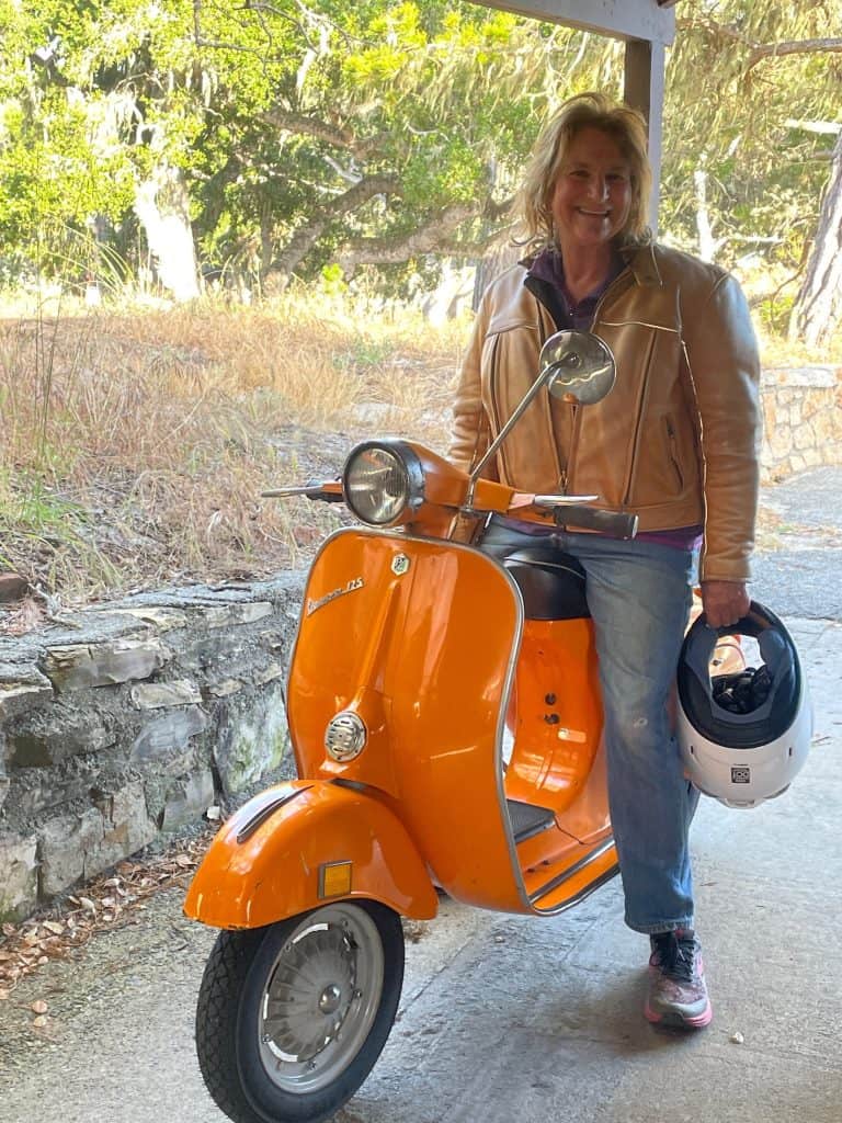 A person in a leather jacket and jeans sits on an orange 1973 Vespa Primavera 125 small frame in Pacific Grove near Monterey, CA, photo ©Kate Dana