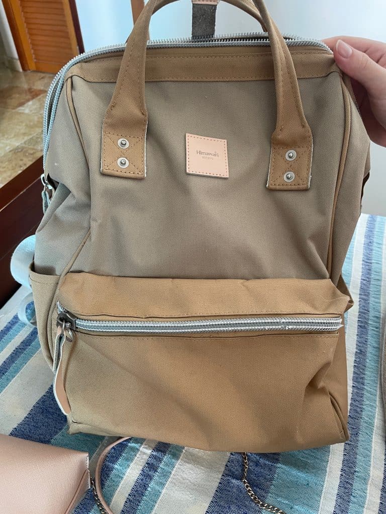 A person's hand shows a tan-colored, medium-sized backpack with zippers, pockets, and handles resting on a striped blue bedspread, photo ©Kate Dana