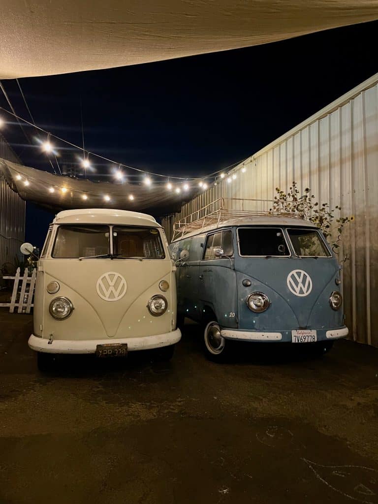 Two vintage Volkswagen buses, one cream colored and one blue, with VW emblems on the front, sit parked under string lights next to a building, photo ©Kate Danaå