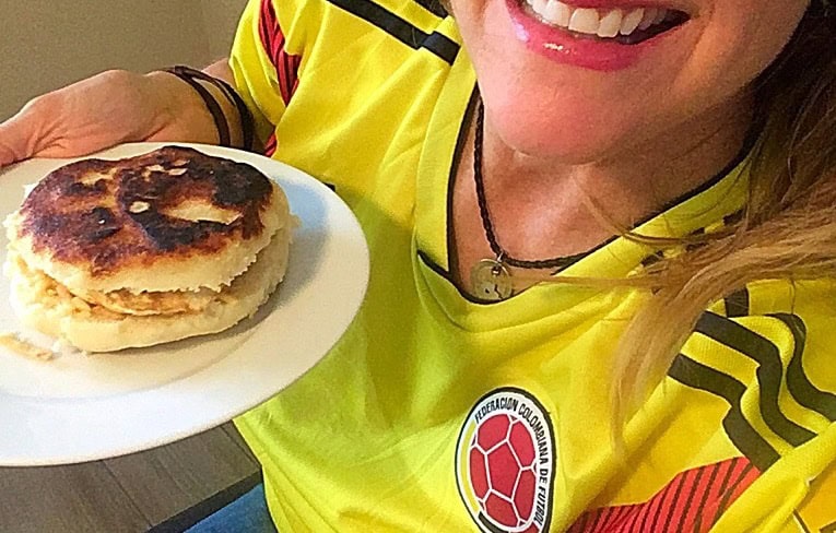 Cocameca founder Kate Dana, wearing a yellow Colombian futbol jersey, holds a golden brown arepa on a plate, photo ©Kate Dana