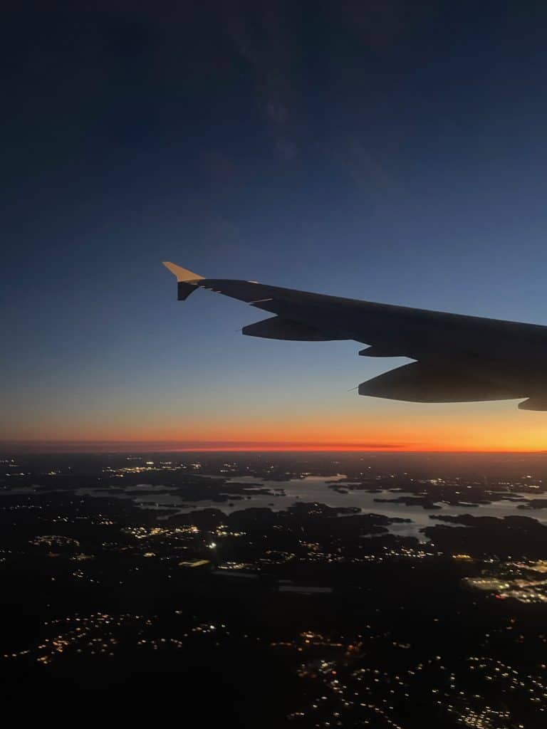 A view from an airplane window shows a wing with an orange sunset horizon in the background, photo ©Kate Dana