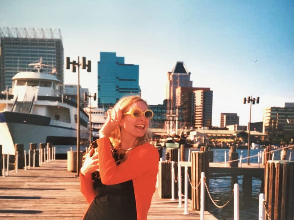 Cocameca founder Kate Dana stands on a pier wearing an orange sweater and large yellow sunglasses in Baltimore,
Maryland, photo ©Kate Dana
