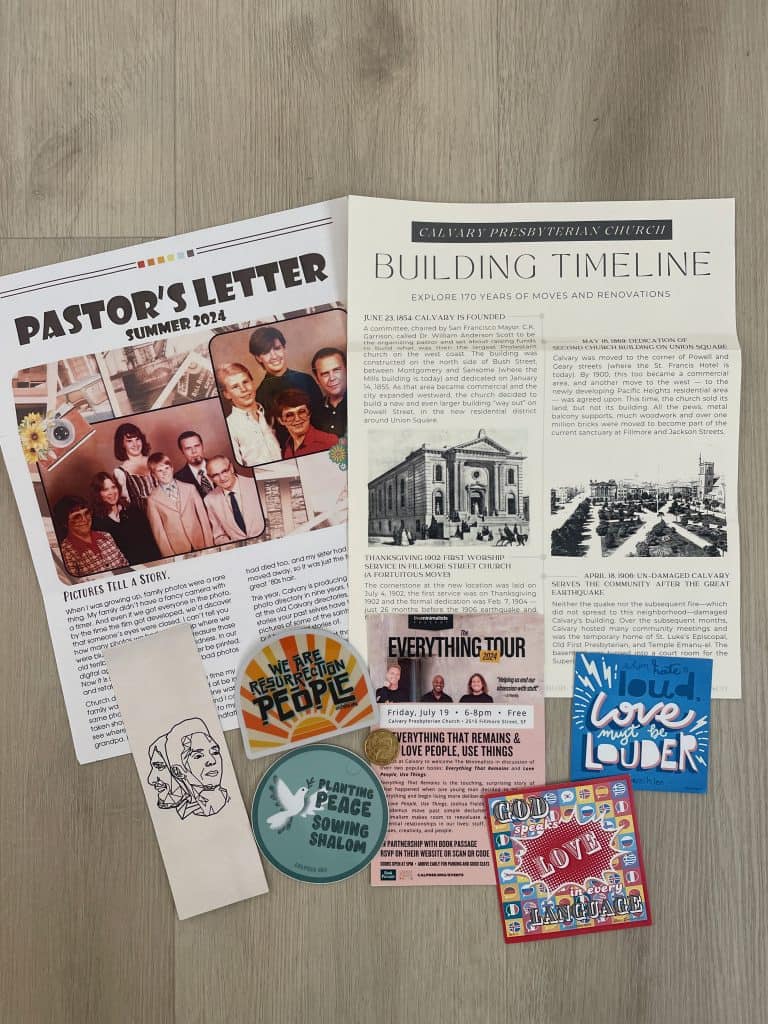A colorful postcard announcing a tour stop date for The Minimalists in San Francisco, CA at Calvary Presbyterian Church, with newsletters and stickers on a wood background, photo ©Kate Dana