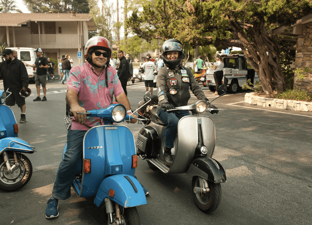 Two people on vintage Vespa scooters smile for the camera under helmets at Vintage Vespa Days 2021 in Los Gatos, CA, photo ©Kate Dana