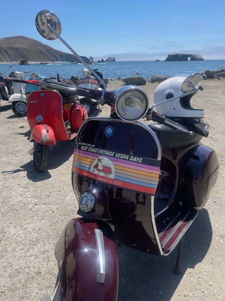 A purple scooter and a red scooter parked close to each other on a beach facing near Santa Rosa, CA for West Coast Vintage Vespa Days 2023, photo ©Kate Dana