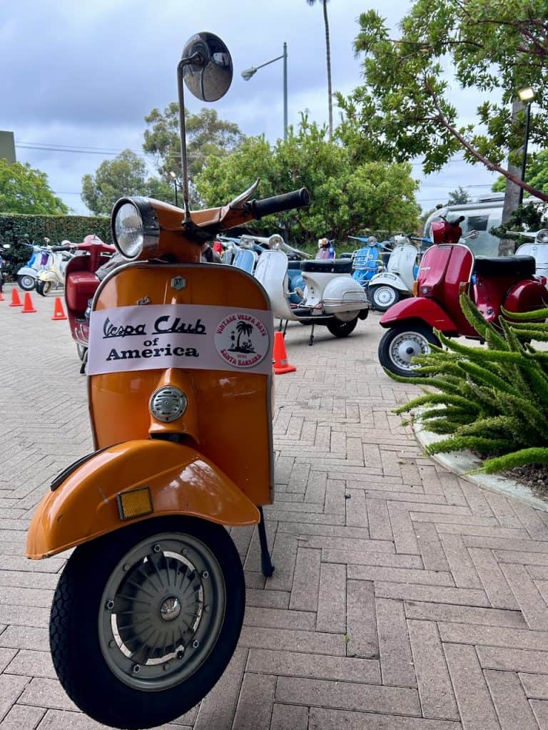 An orange Vespa small frame in front of A group of colorful vintage scooters outside the Leta Hotel in Goleta, CA for West Coast Vintage Vespa Days, photo ©2024 Kate Dana