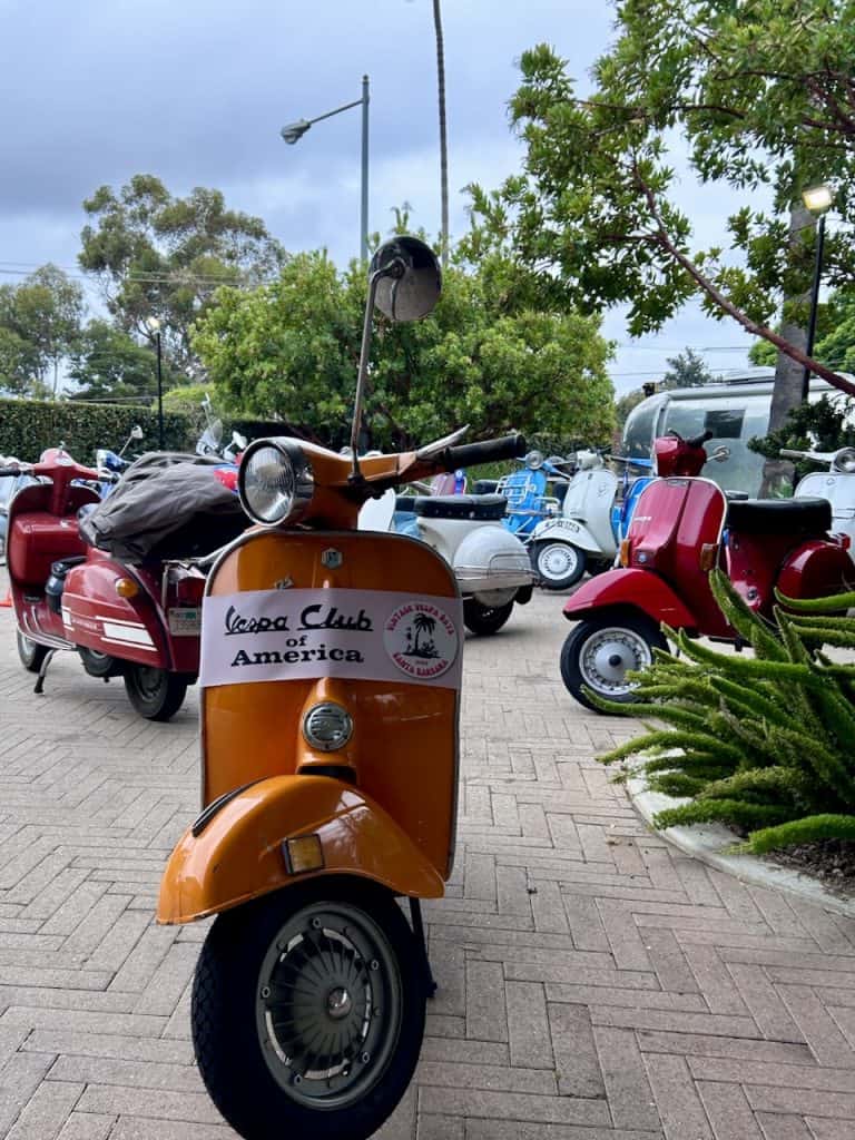 An orange Vespa small frame in front of A group of colorful vintage scooters outside the Leta Hotel in Goleta, CA for West Coast Vintage Vespa Days, photo ©2024 Kate Dana