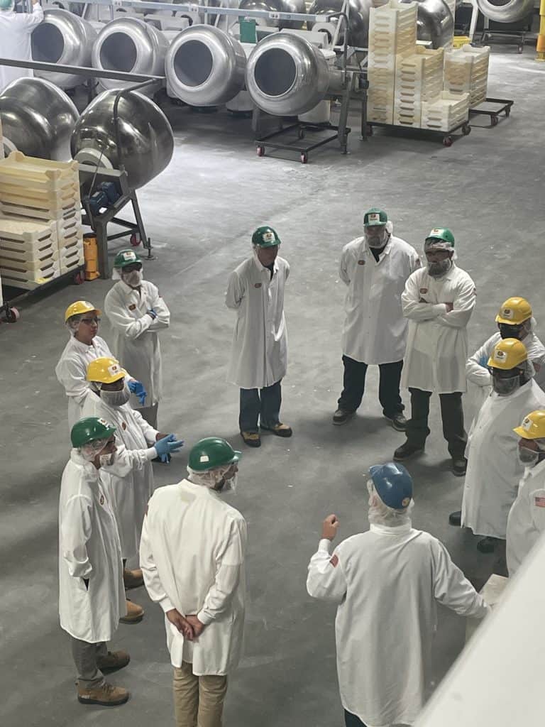 A group of workers dressed in white lab coats, wearing protective eye goggles and hard hats, participate in a group meeting inside the Jelly Belly Factory in Fairfield, CA, photo ©Kate Dana