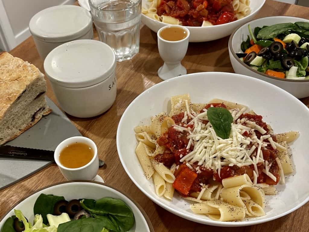 Hearty bowls of pasta with red sauce, cheese, and fresh basil on a table with colorful green salad and small cups of dressing, photo ©Kate Dana
