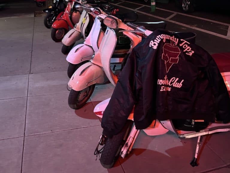 A Burgundy Topz club jacket draped a scooter next to other scooters on a sidewalk, photo ©Kate Dana