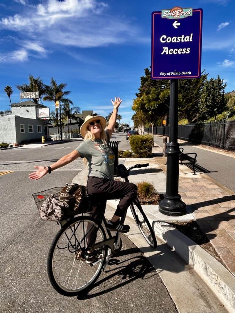 A person wearing a hat and sunglasses stops on a bicycle next to a Coastal Access sign near Shell Beach, California