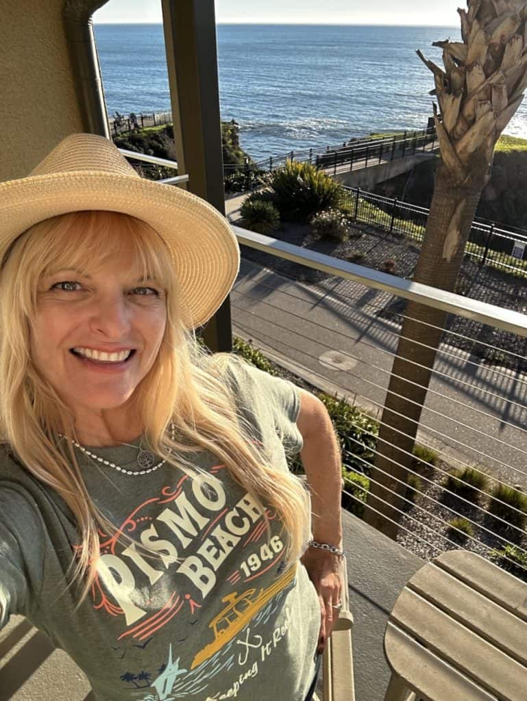 Kate Dana, founder of Cocameca, smiles wearing a hat and Pismo Beach t-shirt on a balcony overlooking the ocean at Inn at the Cove near Pismo Beach, California