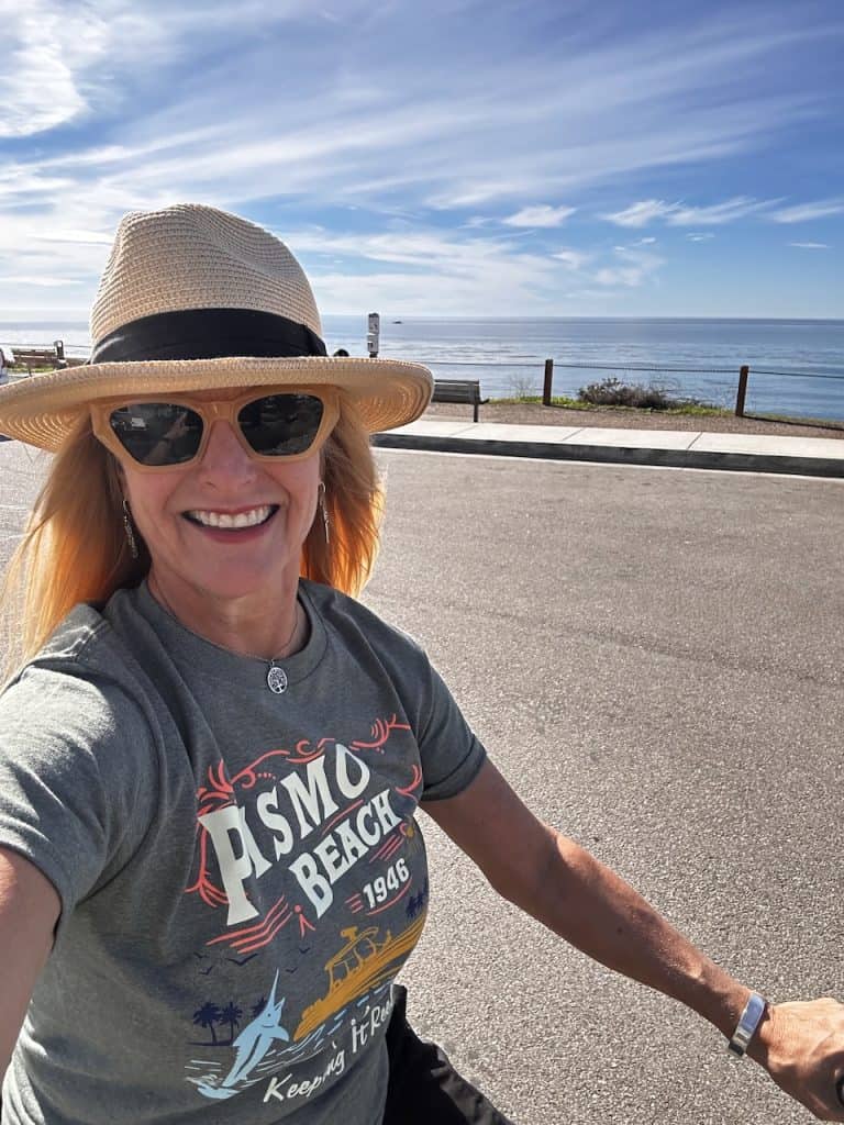 A smiling person in a hat and sunglasses cycles near the ocean wearing a Pismo Beach t-shirt