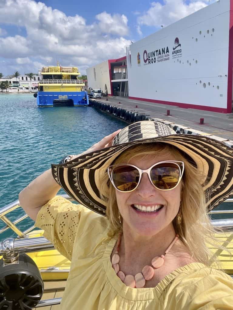 Kate Dana, founder of Cocameca, smiles at the camera wearing a yellow blouse, sunglasses, and a hat near a ferry in Isla Cozumel, Mexico, ©Kate Dana