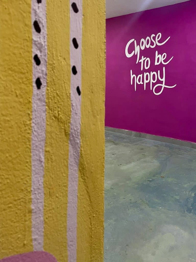 A wall features a pink and yellow painted surface and the words "Choose to be Happy" on Isla Cozumel, Mexico, photo ©Kate Dana