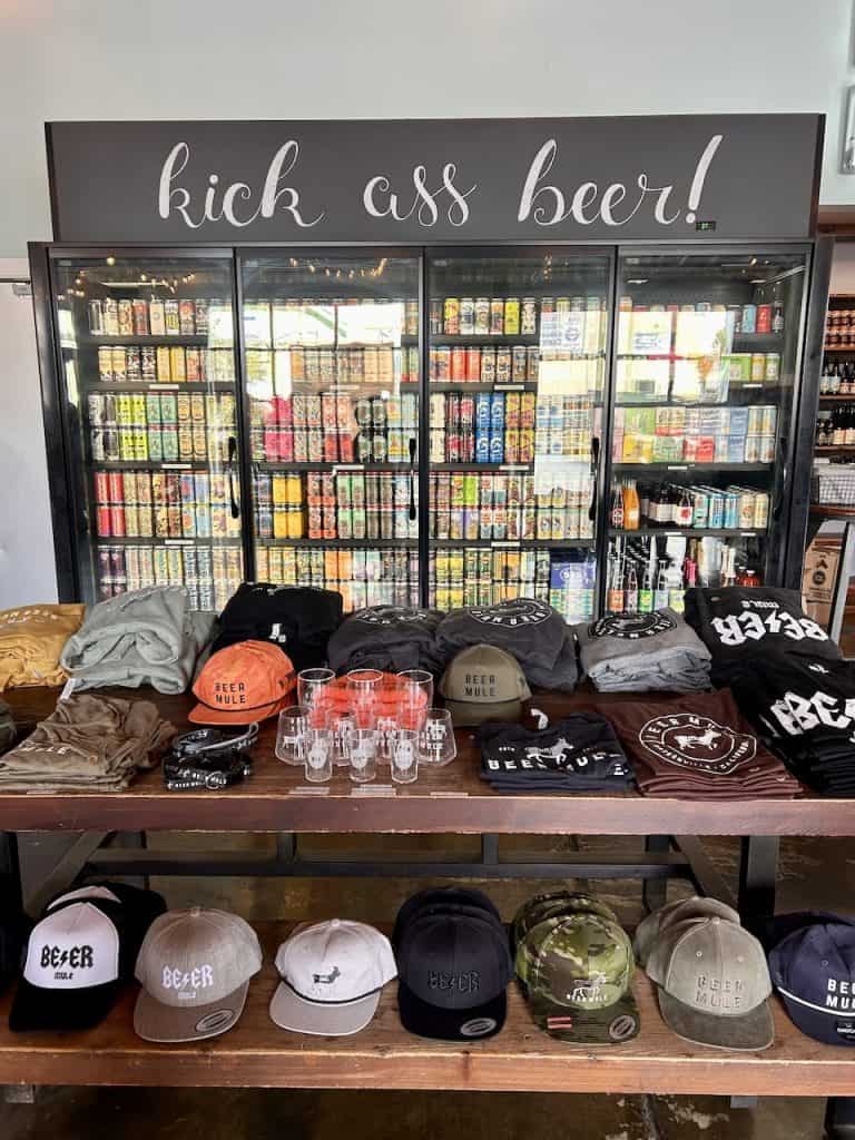 A table of merchandise in front of a stocked refrigerator case at Beer Mule inside The Hangar in Watsonville, CA, photo ©Kate Dana