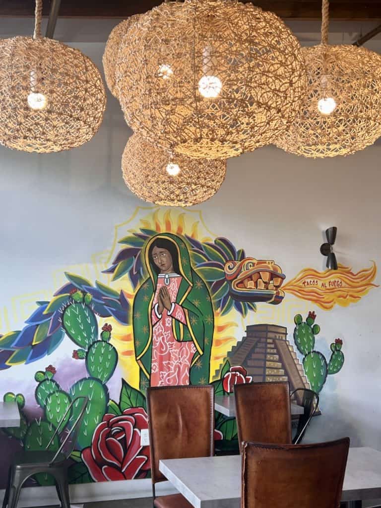 A wall with a mural of the Virgin de Guadalupe lighted below rattan swag lamps in Tacos al Fuego at the Hangar in Watsonville, CA, photo ©Kate Dana