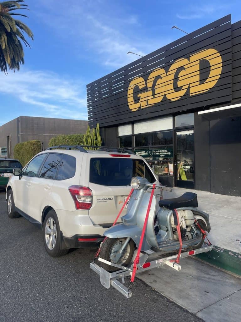 A parked, white car carries a vintage Lambretta on a rack in front of a store reading "Good," photo ©Kate Dana