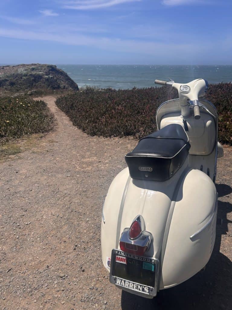 A white vintage Vespa scooter parked facing the ocean on a scenic cliff near Jenner,  California, photo ©Kate Dana