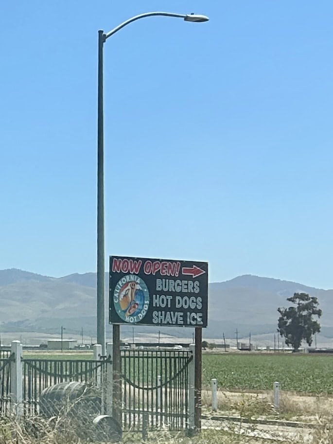 A colorful, hand painted sign in a park offers burgers, hot dogs, and shave ice, photo ©Kate Dana